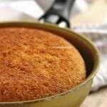 Frigideira pequena de 13 cm dourada com bolo de fubá dentro, na foto aparece apenas metade da frigideira enquadrada, a frigideira está sobre uma tábua de madeira e ao fundo um tecido bege e branco quadriculado