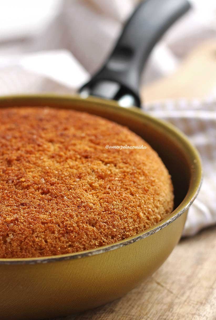 Frigideira pequena de 13 cm dourada com bolo de fubá dentro, na foto aparece apenas metade da frigideira enquadrada, a frigideira está sobre uma tábua de madeira e ao fundo um tecido bege e branco quadriculado