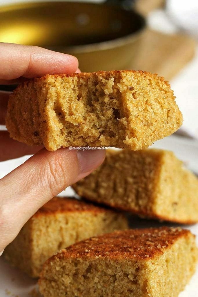 Mão segurando pedaço mordido de bolo de fubá feito na frigideira, abaixo os outros 3 pedaços de bolo amontoados e desfocados