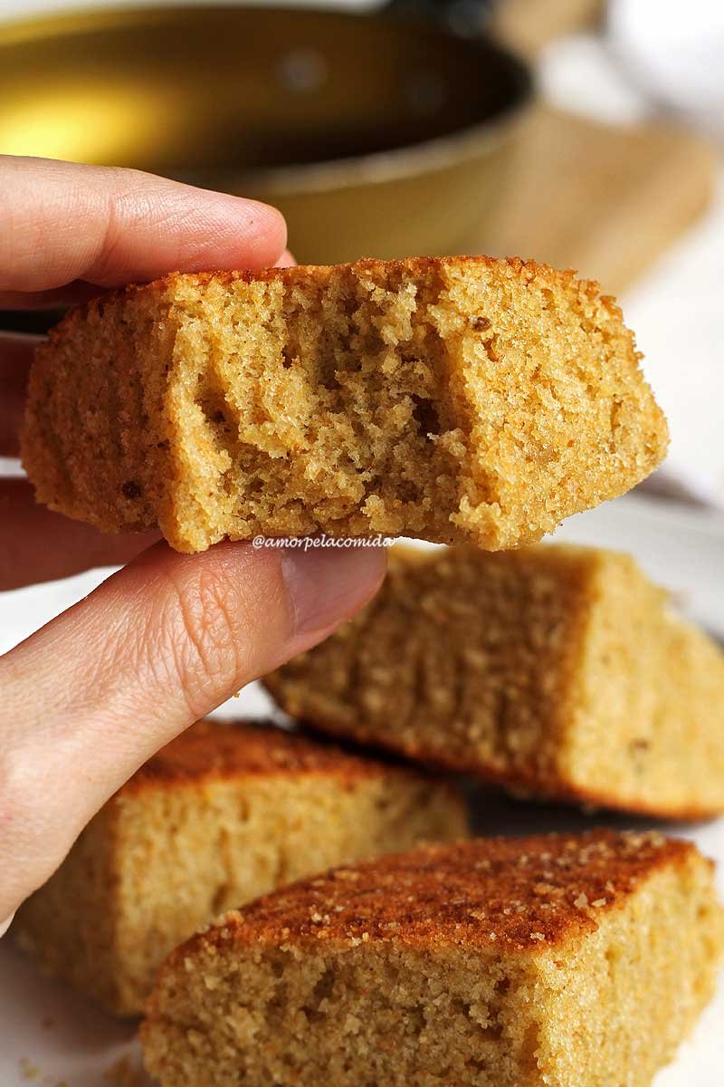 Mão segurando pedaço mordido de bolo de fubá feito na frigideira, abaixo os outros 3 pedaços de bolo amontoados e desfocados