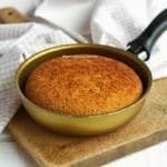 Frigideira pequena de 13 cm dourada com bolo de fubá dentro, a frigideira está sobre uma tábua de madeira e ao fundo um tecido bege e branco quadriculado