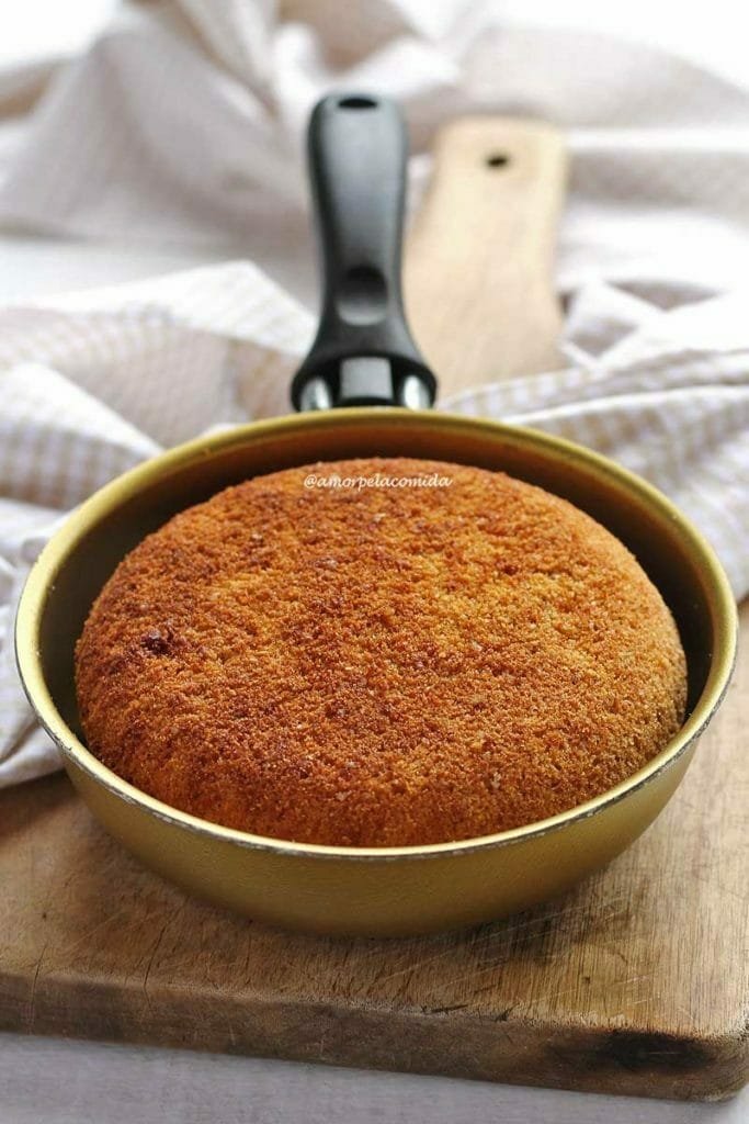 Frigideira pequena de 13 cm dourada com bolo de fubá dentro, a frigideira está sobre uma tábua de madeira e ao fundo um tecido bege e branco quadriculado