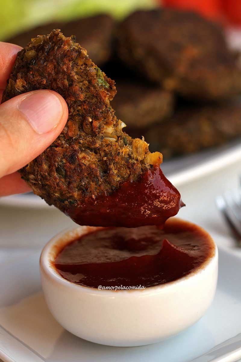 Mão segurando hambúrguer de ervilha partido sendo mergulhado no ketchup, o ketchup está em um pote branco pequeno