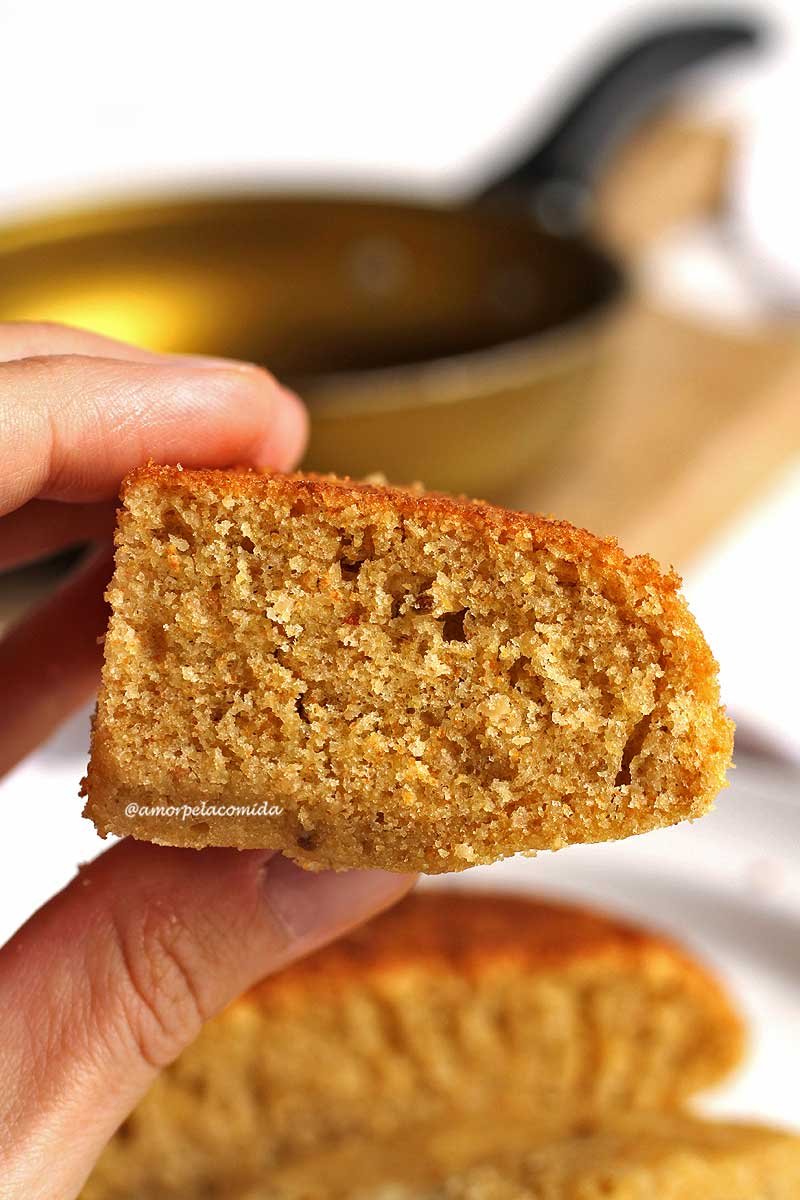 Mão segurando 1/4 do bolo de frigideira pequeno de 13 cm, abaixo os outros 3 pedaços e ao fundo a frigideira dourada vazia sobre uma tábua de madeira desfocado