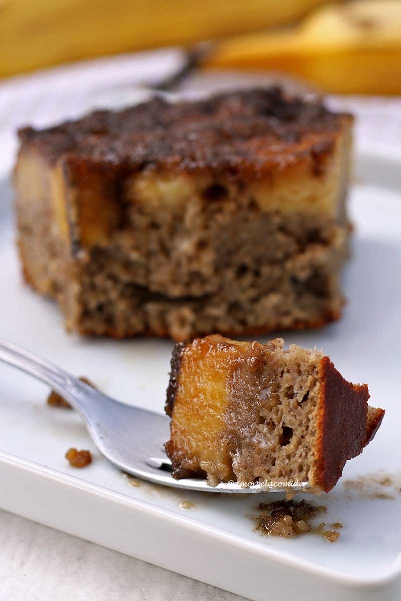 Garfo apoiado em um prato branco com uma garfada de bolo de banana caramelizada, ao fundo o restante da fatia desfocada