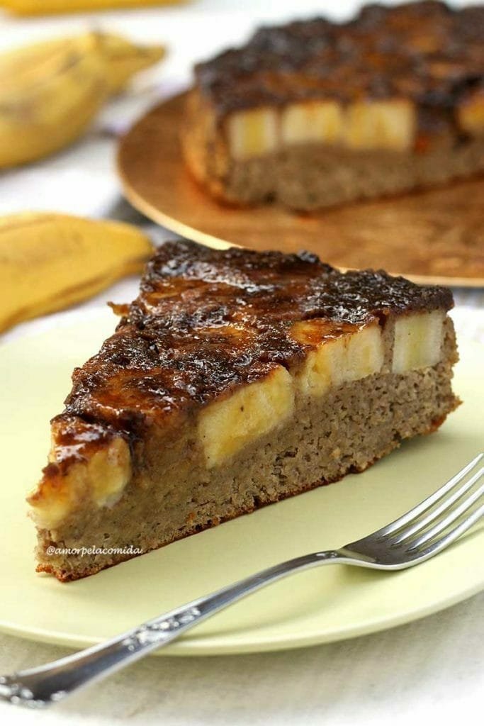 Fatia triangulas de bolo de banana com cobertura de banana caramelizada, ao fundo o bolo partido e algumas bananas em uma mesa branca