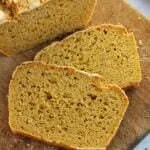 Duas fatias de pão de milho sobrepostas na diagonal sobre uma tábua de madeira, ao lado o pão cortado visto de cima em uma mesa branca