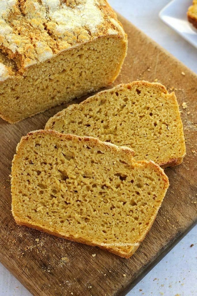 Duas fatias de pão de milho sobrepostas na diagonal sobre uma tábua de madeira, ao lado o pão cortado visto de cima em uma mesa branca