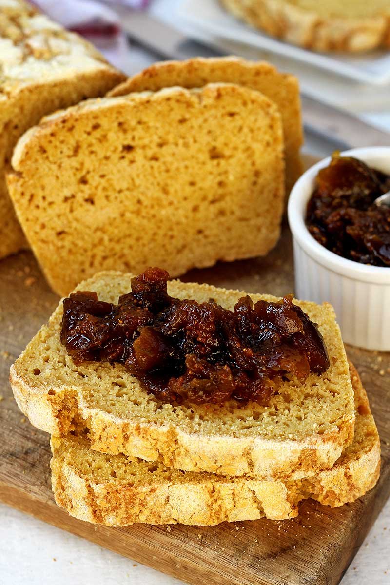 Duas fatias sobrepostas em uma tábua de madeira, a fatia de cima tem geleia de figo em pedaços, ao fundo mais duas fatias cortadas apoiadas no pão de milho, ao lado um pote branco pequeno com geleia de figo