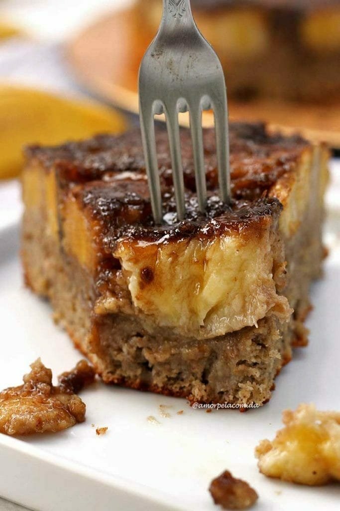 Fatia de bolo com bananas caramelizadas no topo, a fatia já está faltando uma garfada e na foto tem um garfo indicando onde será a próxima garfada