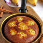 Bolo de banana com rodelas de banana na superfície dentro de uma frigideira pequena dourada sobre tábua de madeira em uma mesa branca com bananas ao fundo desfocadas