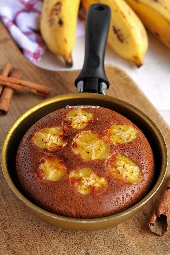 Bolo de banana com rodelas de banana na superfície dentro de uma frigideira pequena dourada sobre tábua de madeira em uma mesa branca com bananas ao fundo desfocadas