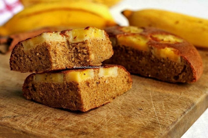 Dois pedaços de bolo de banana sobreposto em uma tábua de madeira, ao fundo metade do bolo de banana de frigideira e mais bananas frescas