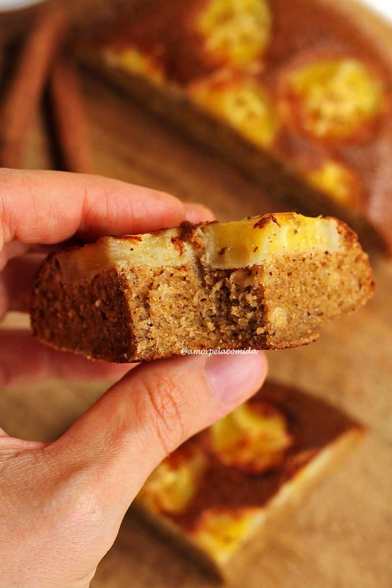 Mão segurando pedaço de bolo de banana mordido, no topo do bolo rodelas de banana abaixo sobre tábua de madeira o resto do bolo de banana