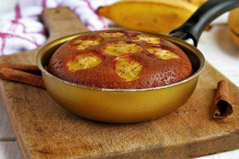 Bolo de banana dentro de uma frigideira dourada com rodelas de banana por cima do bolo em uma tábua de madeira sobre uma mesa branca com bananas desfocadas ao fundo