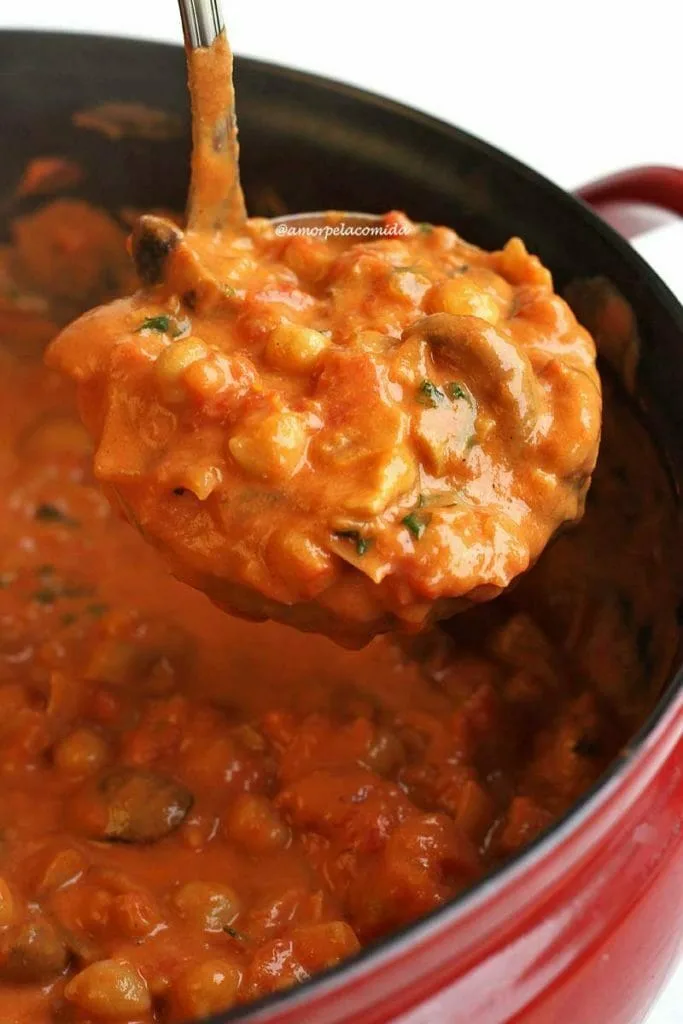 Cucharón lleno de stroganoff de champiñones con garbanzos en la sartén, la sartén tiene un interior negro y el exterior es rojo