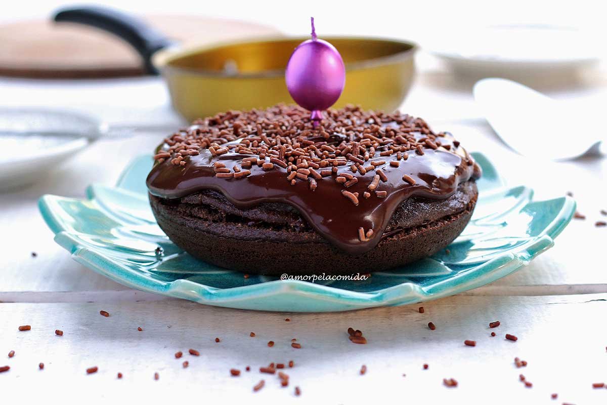 Bolo de chocolate redondo pequeno sobre prato azul turquesa, ao fundo uma frigideira amarela desfocada