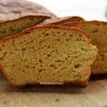 Pão caseiro fatiado, são três fatias apoiadas no pão sobre uma tábua de madeira em uma mesa branca