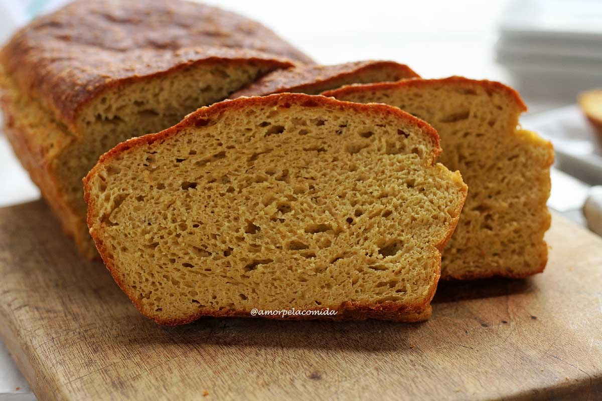 Pão caseiro fatiado, são três fatias apoiadas no pão sobre uma tábua de madeira em uma mesa branca