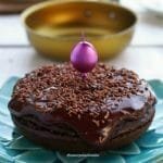 Bolo de chocolate redondo pequeno sobre prato azul turquesa, ao fundo uma frigideira amarela desfocada