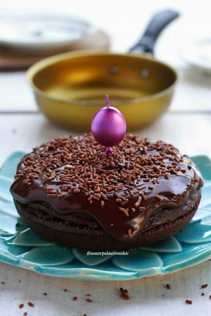 Bolo de chocolate redondo pequeno sobre prato azul turquesa, ao fundo uma frigideira amarela desfocada