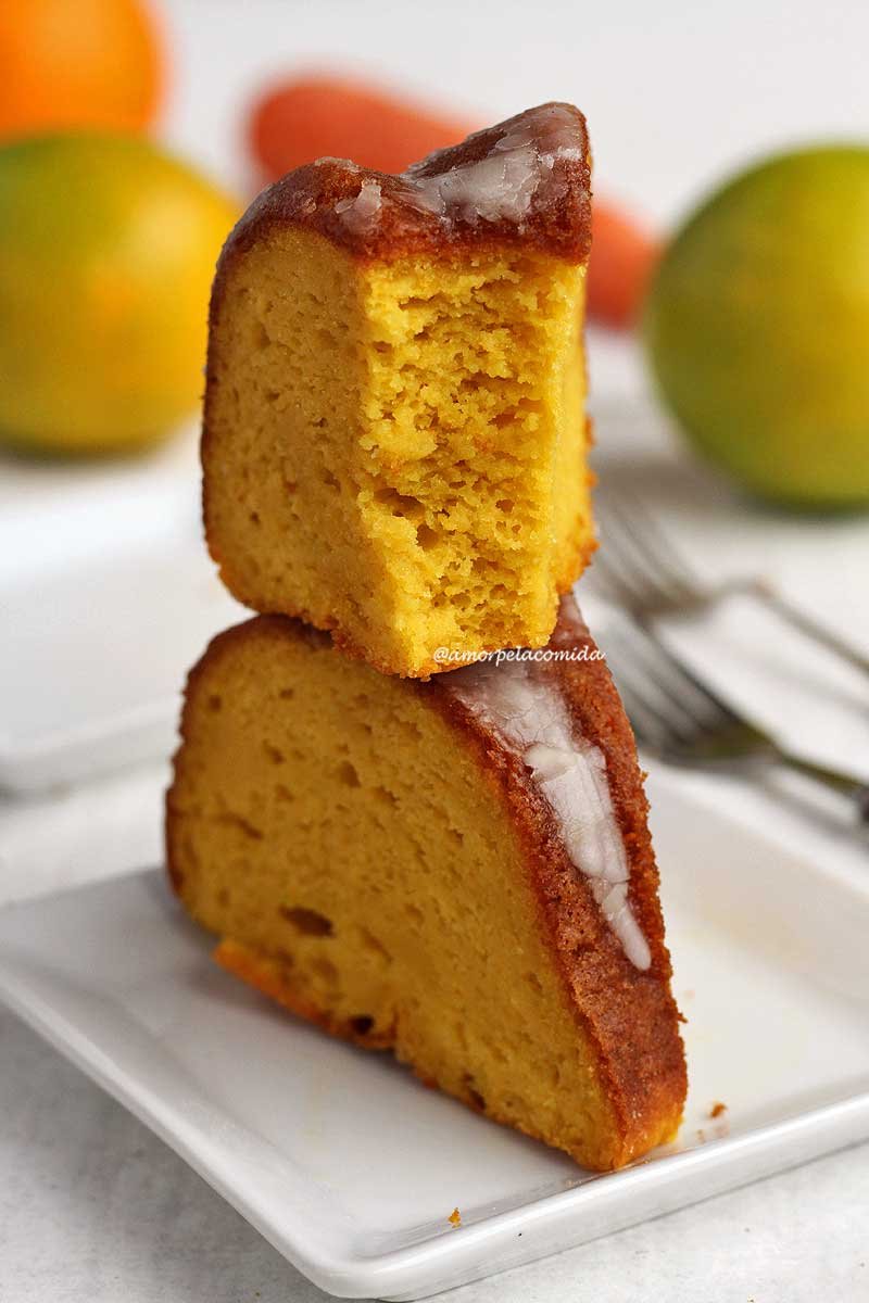 Duas fatias de bolo de cenoura com laranja sobrepostas em um prato branco quadrado pequeno, a fatia de cima está mordida