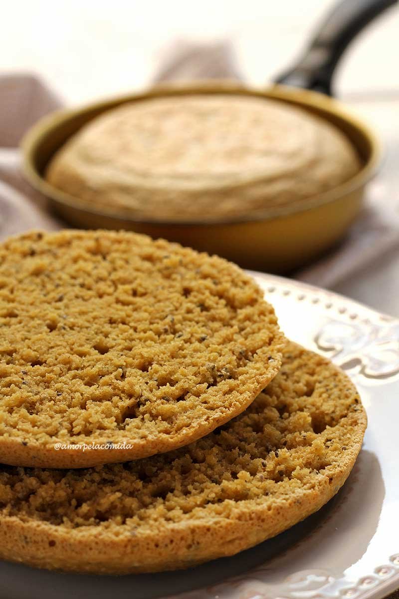 Pão redondo pequeno cortado ao meio mostrando a textura aerada da fatia sobre prato branco, ao fundo a frigideira com um pãozinho dentro desfocado da cena