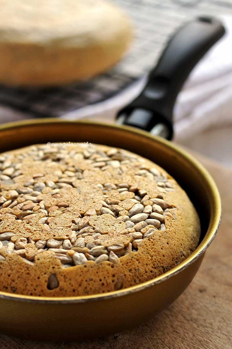 Pão redondo pequeno dentro de uma frigideira dourada sobre uma tábua de madeira em uma mesa branca, na superfície do pão tem várias sementinhas de girassol decorando o pão