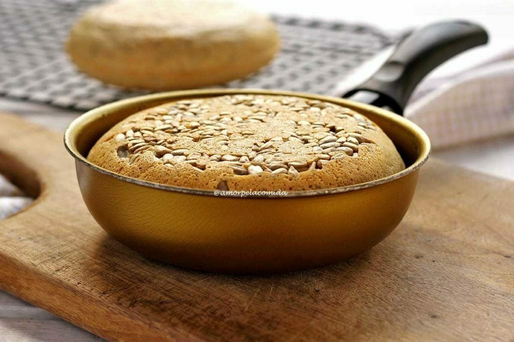 Pão pequeno feito dentro de uma frigideira dourada, no topo do pão sementes de girassol. A frigideira está sobre uma tábua de madeira e ao fundo tem uma grelha preta com mais um pão redondo pequeno de frigideira desfocado da cena principal