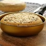 Pão pequeno feito dentro de uma frigideira dourada, no topo do pão sementes de girassol. A frigideira está sobre uma tábua de madeira e ao fundo tem uma grelha preta com mais um pão redondo pequeno de frigideira desfocado da cena principal