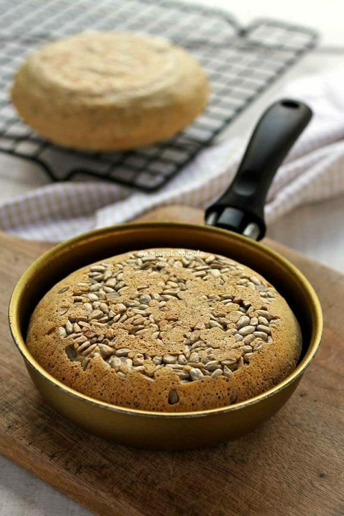 Pão pequeno dentro de uma frigideira dourada de 13 cm sobre uma tábua de madeira, ao fundo tem um pão de frigideira esfriando sobre uma grade preta