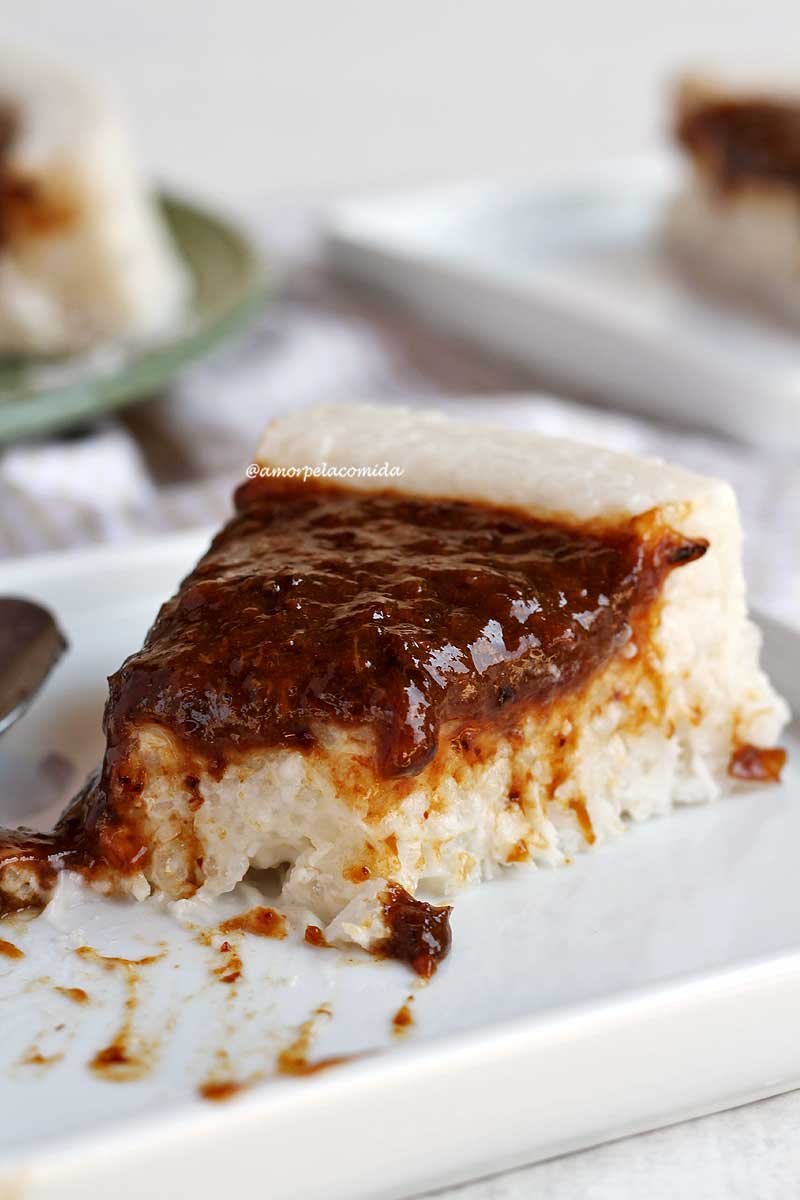 Pedaço de bolo de tapioca com coco e calda de ameixa já faltando uma colherada sobre um prato branco quadrado