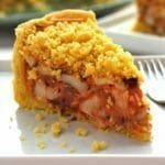 Fatia de empadão de palmito sobre prato quadrado branco, na foto aparece o recheio com bastante palmito, molho de tomate e azeitonas, na cobertura uma farofinha crocante