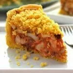Fatia de empadão de palmito sobre prato quadrado branco, na foto aparece o recheio com bastante palmito, molho de tomate e azeitonas, na cobertura uma farofinha crocante