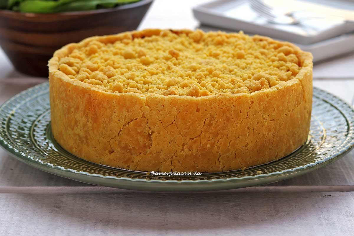 Empadão redondo com casquinha crocante e farofinha crocante por cima sobre um prato verde em uma mesa branca