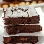 Três fatias de brownie de chocolate empilhadas sobre prato quadrado pequeno, as fatias estão separadas por papel manteiga