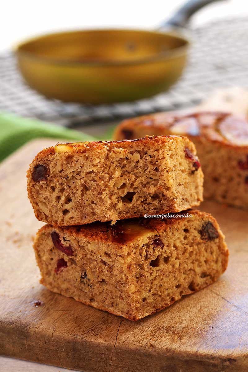 Dois pedaços de bolo de maçã sobrepostos em uma tábua de madeira de cor clara, o pedaço de cima está mordido mostrando a textura aerada da massa