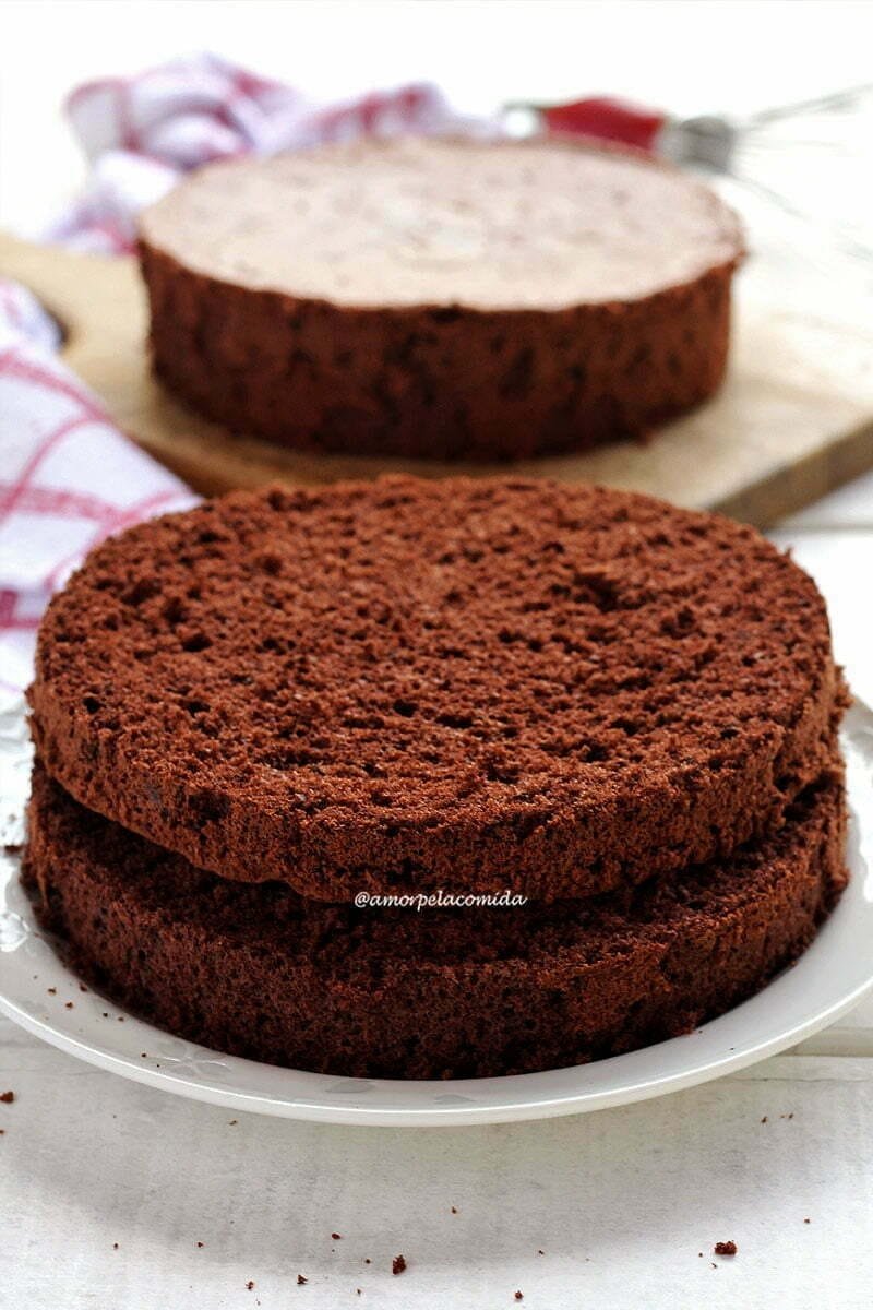 Pão de ló de chocolate cortado ao meio sobre prato branco, ao fundo outra massa de pão de ló sobre tábua de madeira