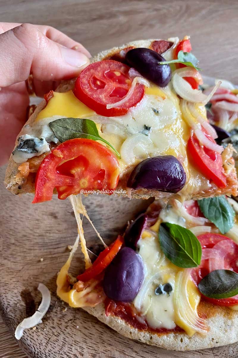 Mão segurando fatia de pizza, abaixo mais dois pedaços de pizza sobre uma tábua de madeira