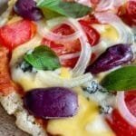 Pizza sobre tábua de madeira, na cobertura queijo, tomate, cebola, manjericão e azeitonas pretas