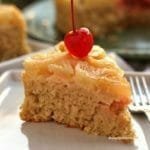 Fatia triangular de bolo de abacaxi com coco, no topo do bolo tem pedaços de abacaxi e uma cereja com cabinho