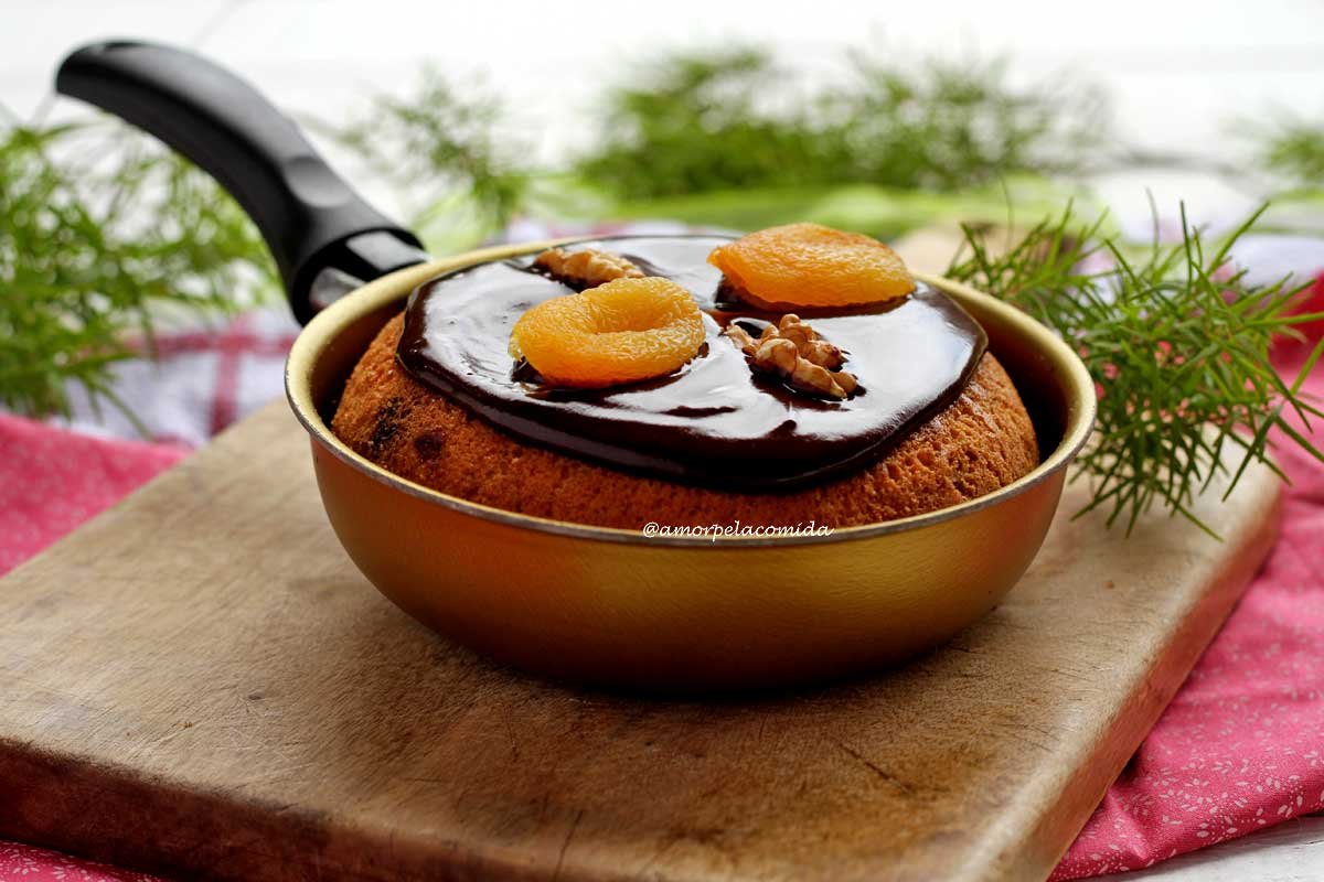 Frigideira pequena dourada sobre tábua de madeira, dentro um panetone decorado com cobertura de chocolate cremosa e brilhante com damasco e nozes