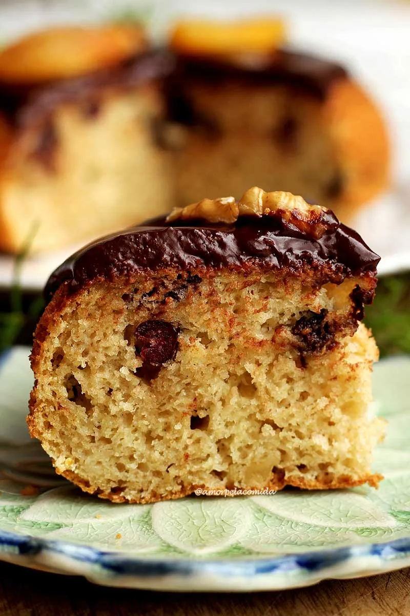Pedaço de panetone com cobertura de chocolate sobre pratinho pequeno, ao fundo o panetone de frigideira desfocado