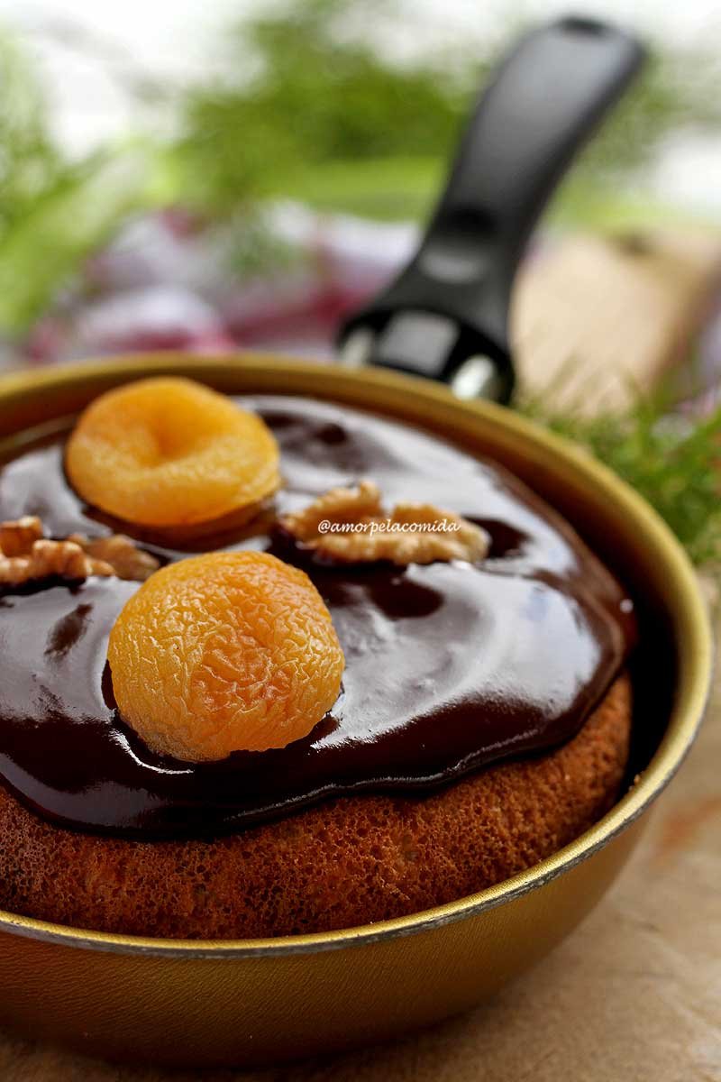 Panetone de frigideira coberto com cobertura cremosa de chocolate, a decoração é com damasco e nozes e a frigideira está sobre uma tábua de madeira de cor clara