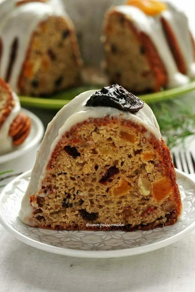 Fatia de bolo de frutas com cobertura branca, sobre a cobertura uma ameixa seca decorando e ao fundo o bolo partido sobre um prato verde