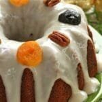 Bolo redondo com furo no meio com cobertura branca e frutas secas decorando o topo sobre um prato verde