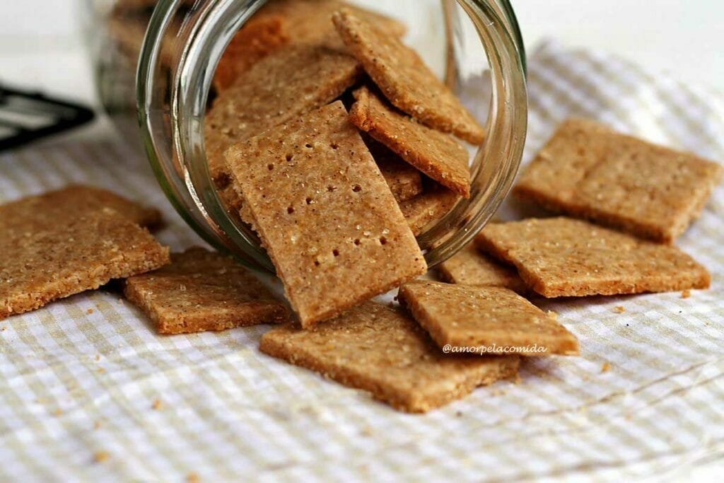 Vários biscoitinhos saindo de um vidro transparente apoiados sobre pano quadriculado branco e bege