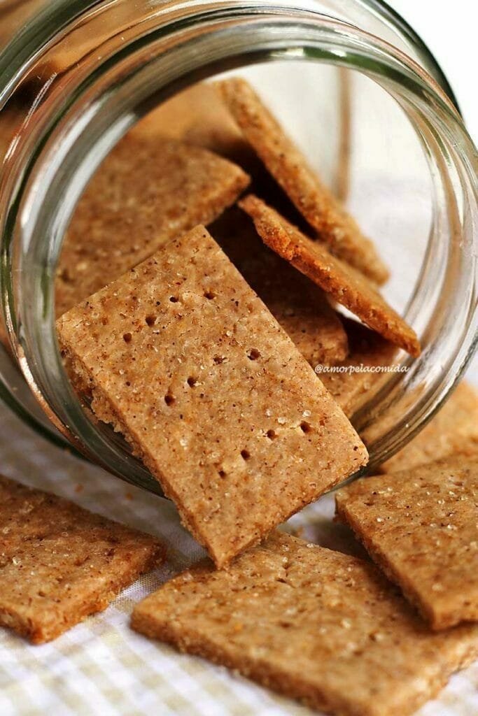 Biscoitinho retangulares saindo de um vidro transparente que está deitado sobre uma mesa branca