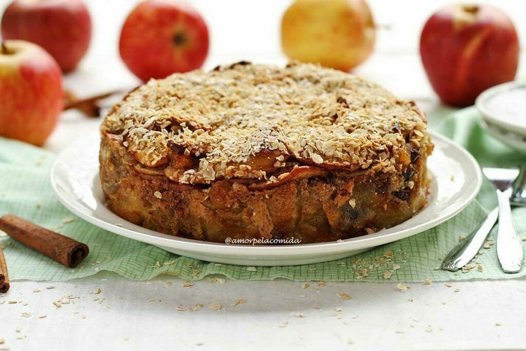 Torta de maçã redonda com cobertura de flocos de aveia sobre prato branco, ao fundo algumas maçãs desfocadas