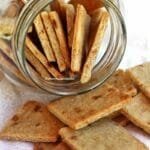 Vários biscoitinhos fininhos e crocantes dentro e fora de um vidro transparente que está deitado em uma mesa branca